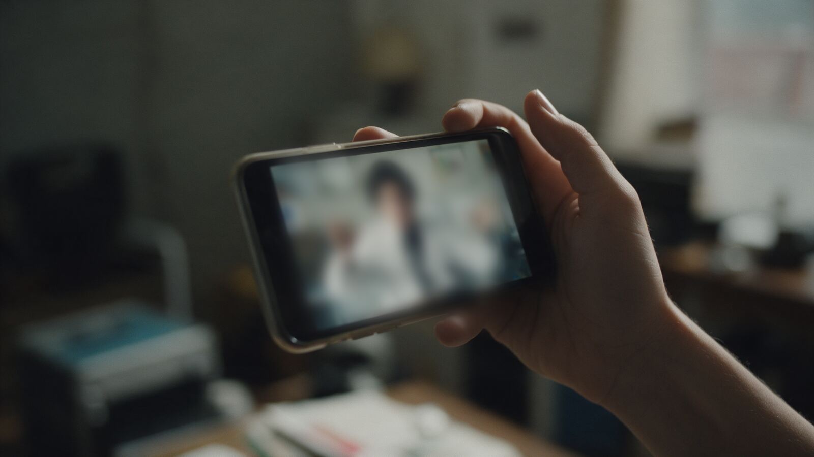Ruka drží telefon s rozmazaným videem člověka v bílém plášti
