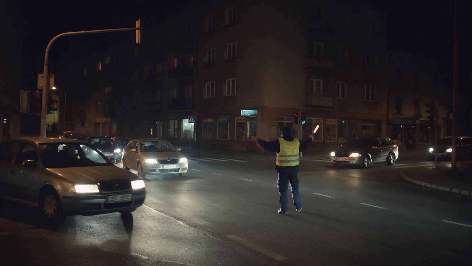 Křižovatka v noci bez proudu, policista zezadu řídí dopravu svítící hůlkou